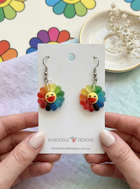 Rainbow flower drop earrings with smiling face centre on Kaboodle Designs card, held in hand