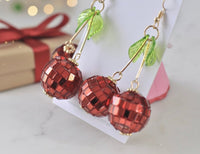 Close-up of mirrorball cherry earrings showing faceted red beads, gold stems and green leaf detail