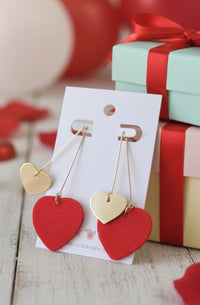 Red and gold heart dangle earrings on a card, displayed against gift boxes and red ribbon