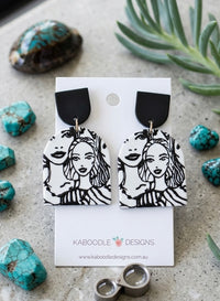 Black and white resin drop earrings with line-art faces of two women, arch-shaped with black stud top, shown on card