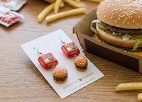 Burger box and burger charm earrings displayed on card beside a real burger and fries
