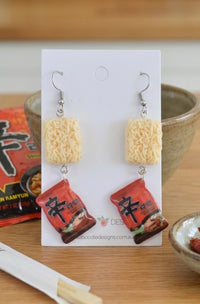 Resin drop earrings shaped like noodle blocks and red Shin Ramyun packets on a display card