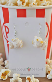 Cream and yellow resin popcorn drop earrings displayed on a card in front of a striped popcorn bucket