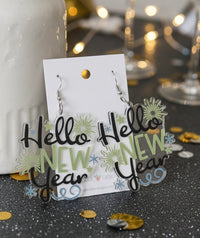 Acrylic 'Hello New Year' drop earrings with firework and star design in black, green and blue, shown on card