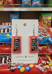 Resin M&M packet dangle earrings in pink packaging on a Kaboodle Designs card, shown against a candy aisle backdrop