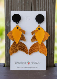 Gold acrylic koi fish drop earrings on black studs, displayed on Kaboodle card against timber background