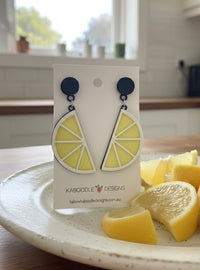 Acrylic lemon slice drop earrings on card with navy studs, shown beside fresh lemon wedges on a plate