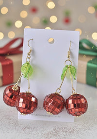 Red faceted disco-ball cherry earrings displayed on white Kaboodle card against Christmas lights