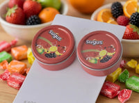 Close-up of two Sugus tin earrings on a card, surrounded by loose wrapped lollies and fruit