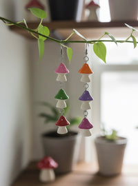 Mushroom drop earrings hanging from a leafy vine, showing pink, orange, green, purple, red and magenta toadstool caps