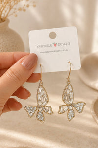 Gold-tone rhinestone butterfly dangle earrings held in hand on Kaboodle Designs card