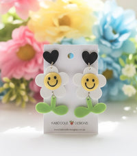 Acrylic daisy smiley face drop earrings with black heart stud and green stem, shown on card