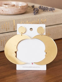 Gold mirrored crescent moon earrings with engraved face on Kaboodle Designs card, resting against books