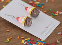 Pair of cupcake earrings on card viewed from an angle, scattered with colourful sprinkles