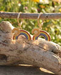 Rainbow sunflower drop earrings hanging from a driftwood branch, showing the striped enamel rainbow and gold sunflower detail