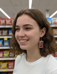 Close-up side view of a model wearing one M&M packet earring, smiling in a supermarket aisle