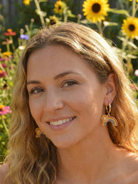 Close-up of a woman wearing a gold hoop earring with rainbow and sunflower charm, smiling in front of sunflowers