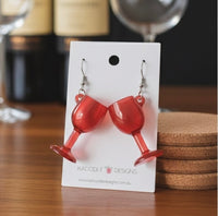 Red wine glass novelty drop earrings on a Kaboodle Designs card, styled with wine bottles and coasters