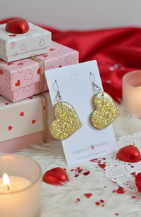Pair of gold glitter heart earrings on card, styled with red heart props and candles