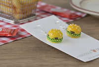Angled view of cheeseburger earrings on display card beside a picnic-themed background