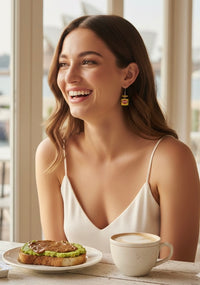 Woman wearing a single Vegemite jar drop earring while smiling, with avocado toast and coffee on the table