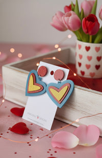Pair of layered heart drop earrings displayed on card beside pink tulips and heart confetti