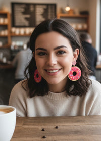 Woman wearing pink donut sprinkle earrings with glitter heart studs, smiling in a cafe