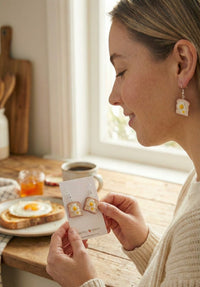 Woman holding a card of egg-on-toast earrings while wearing one, breakfast scene with toast and coffee in background