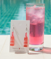 Pair of resin raspberry Vodka Cruiser bottle earrings on card beside a pink poolside drink