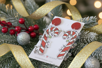 Close-up angled view of candy cane Christmas lights earrings on card, styled with gold ribbon and berries