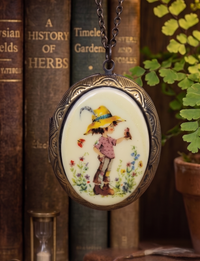 Oval locket necklace with illustrated meadow scene of a girl in a yellow hat holding a bird, shown against vintage books