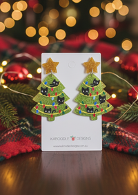 Green glitter Christmas tree drop earrings with black cat faces and gold star studs, on card against fairy lights