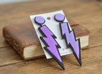 Purple lightning bolt drop earrings on card leaning against a leather book on wooden table