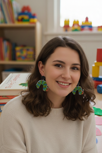 Woman wearing green and blue glitter caterpillar dangle earrings, smiling in a playroom setting