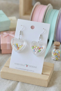 Clear heart-shaped resin earrings filled with pastel hundreds and thousands sprinkles, on card