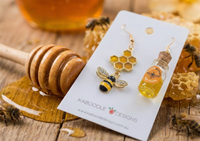 Close-up of honeycomb and jewelled bee charm earring next to honey jar earring with cork lid, styled with honey dipper