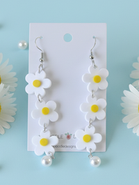 White acrylic daisy dangle earrings with yellow centres and faux pearl drop, shown on card