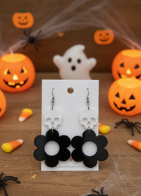 White skull and black flower acrylic drop earrings on a Kaboodle display card, Halloween props behind