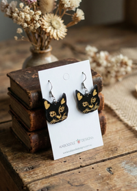 Close-up of black perspex cat face earrings showing gold whisker and eye detail