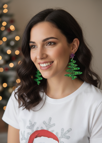 Woman wearing green glitter Christmas tree earrings with star stud, festive lights in background