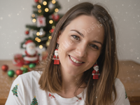 Close-up of a woman wearing red Santa figure earrings in front of a Christmas tree