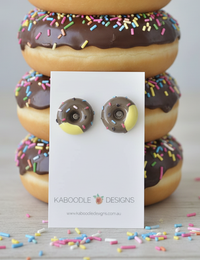 Chocolate donut resin stud earrings with rainbow sprinkles on white card, in front of stacked real donuts