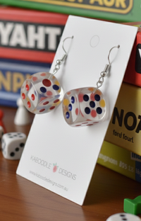 Pair of clear dice earrings displayed against stacked board game boxes