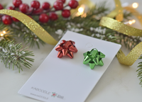Close-up angled view of red and green bow earrings on card, resting among pine branches and gold ribbon