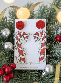 Red glitter stud and candy cane charm earrings with fairy lights design, laid on card among pine and baubles