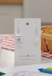 Resin milk carton drop earring with cow mascot print and purple stripe detail, shown on display card beside a matching milk