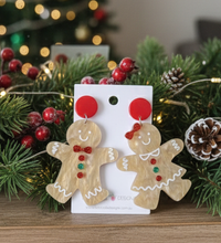 Gingerbread man and woman acrylic drop earrings with red circle tops on card, styled among Christmas greenery
