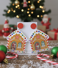 Pair of gingerbread house acrylic earrings with red stud tops on white card, candy canes and baubles nearby