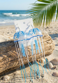 Pair of clear acrylic jellyfish dangle earrings with blue spotted domes, resting against driftwood on a beach