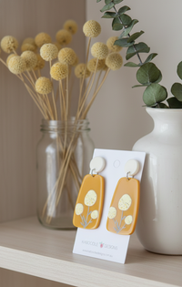 Mustard flower drop earrings displayed on shelf beside dried billy buttons and eucalyptus